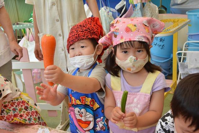 エプロンにマスクをして、にんじんやオクラを持っている園児たちの写真
