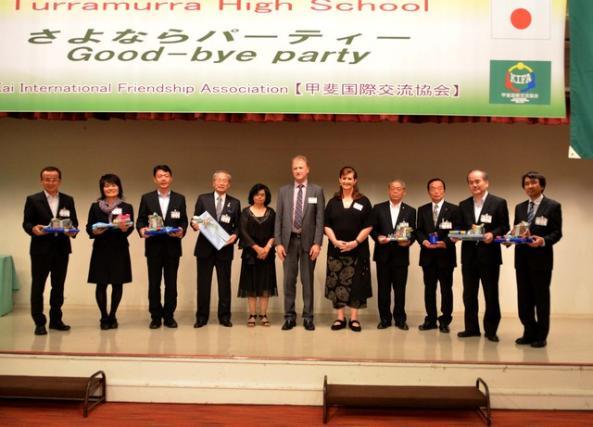 甲斐市や各学校の関係者が記念品を持ち舞台の上で並んでいる写真