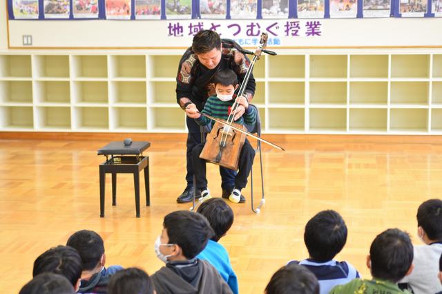 演奏家の男性に支えられながら馬頭琴を持たせてもらう児童の写真