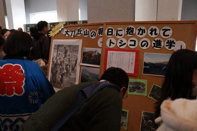 「大刀丘山の朝日に抱かれて~トシコの進学」と書かれたボードに貼られている写真を沢山の人が見ている写真
