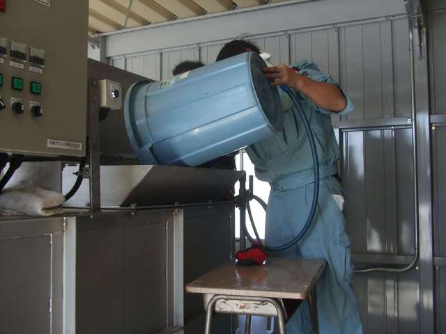 職員が水色のポリバケツを傾け、給食残渣を液肥化する機械に投入している写真