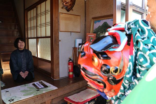 法被を着た男性が住民の女性の前で獅子舞を披露している写真