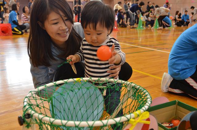 小さな子が母親に手伝ってもらいオレンジ色のボールをカゴに入れている写真