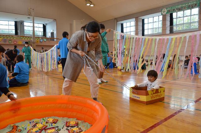 子どもを乗せた箱を母親が引っ張り、カラーテープのすだれの下を通っている写真