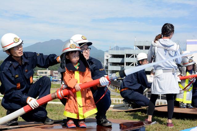 消防士2人の支えられ、消防士の制服を着た小さな男の子は放水の体験をしている写真