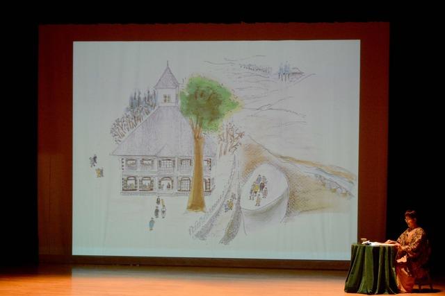 舞台の上で藤巻さんが睦沢小学校の絵のスライドと合わせ話をしている写真