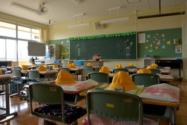 各机に黄色い帽子が置かれた誰もいない教室の写真