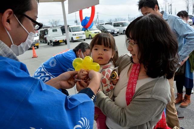 母親に抱かれた小さな女の子が法被を着た関係者から黄色のバルーンアートを受け取っている写真