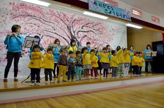 後ろの大人が吹奏楽を演奏し、前の列の子ども達が歌っている写真