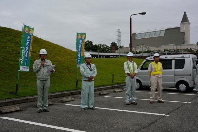 駐車場で4人の作業員が立ち、1人がマイクを持って話している写真