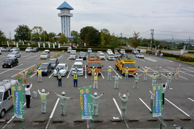 駐車場で約20人の作業員が両手を挙げて甲州弁ラジオ体操をしている写真