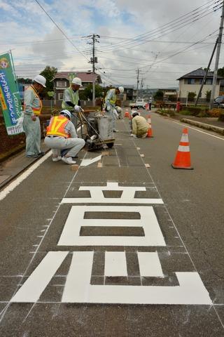 複数の作業員で地面に書かれた「注意」という字に白い路面標示剤を塗っている写真
