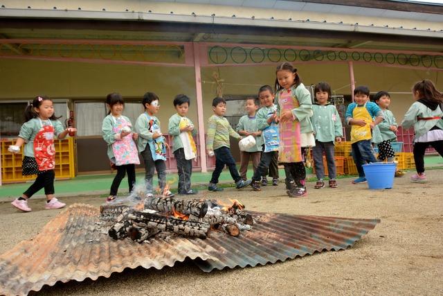 エプロン姿の園児たちがサツマイモを焚き火の中に入れている写真