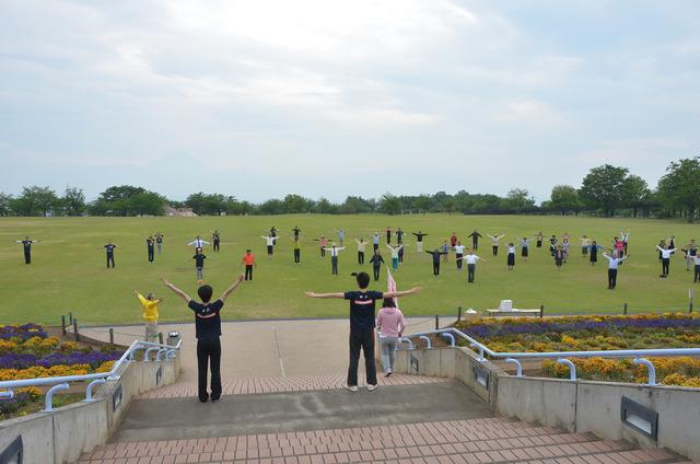 広い芝生の上で沢山の人が大きく手を広げてラジオ体操をしている写真