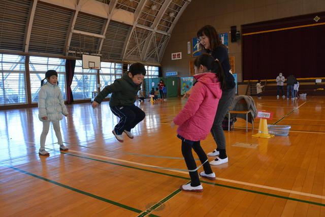 体育館で足にゴムを通して向かい合った女の子二人と、その間で飛ぶ男の子の写真