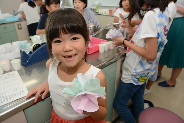 講座「キッチンで科学しよう」にて着色したコーヒーフィルター製の花を持つ正面を向いている女子小学生と着色中の女子小学生たちの写真