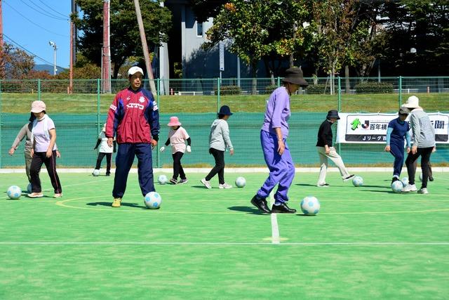 ヴァンフォーレ甲府のコーチの指導のもと参加者たちがサッカーボールをドリブルしている写真