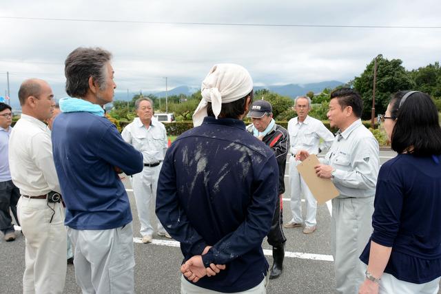 駐車場で作業服をきた男性が集まり、1人の男性が指示を出している写真