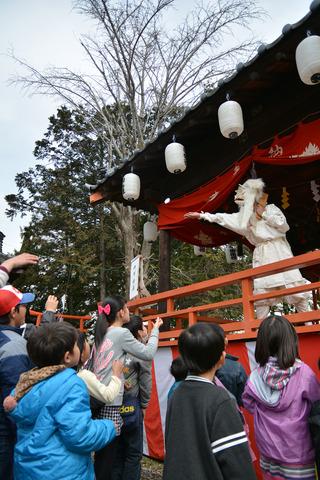 衣装を着て面をつけて舞台からお菓子を投げている写真