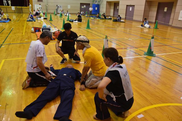 体育館内で倒れている人の周りに4人の男女が集まっている写真