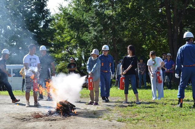 八幡諏訪神社でヘルメットを被った女性が消防団員の指導を受けて火に向かって消火器を使用している写真