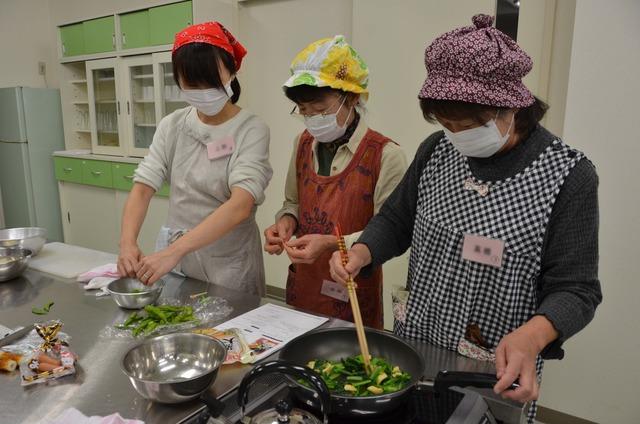 野菜を炒めている1人の女性と枝豆の処理をしている2人の女性の写真