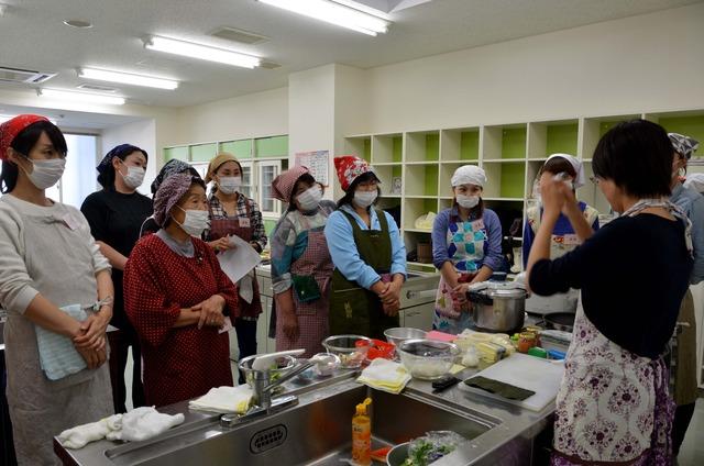 エプロンとマスクを着用した多くの参加者の女性が講師の早川亜希子さんの話を聞いている写真