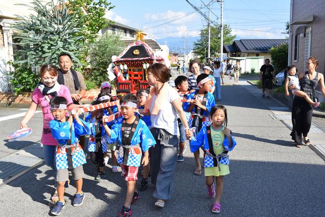 青い法被を着てハチマキをした子どもたちが保護者に付き添われて神輿を担いでいる写真
