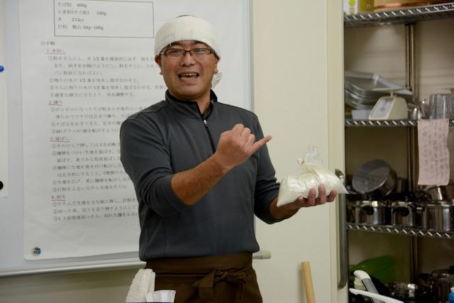 講師の中村まさしさんが袋に入ったソバ粉を持って説明をしている写真