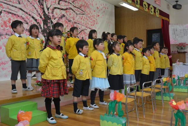 手形で彩られた桜の絵をバックに並んで歌を歌う園児たちの写真