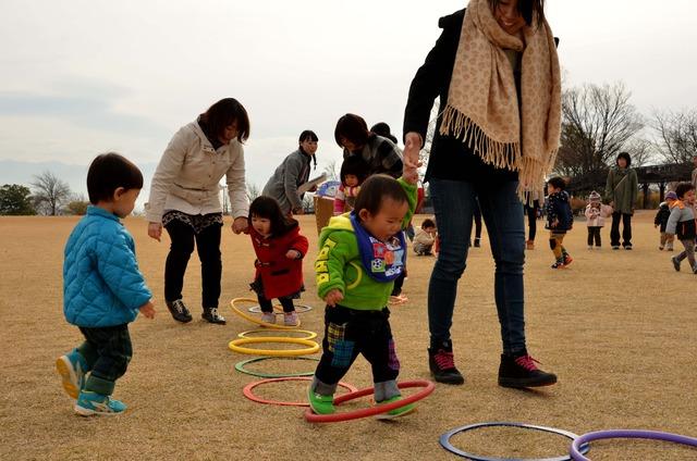 母親に手を引かれた子どもたちが地面に置かれた沢山の輪の中を歩いている写真