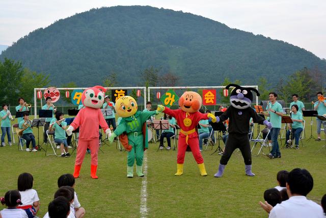 敷島総合公園の芝生の上で、甲斐市敷島吹奏楽団が演奏する前でアンパンマンと仲間のキャラクターが踊っている写真