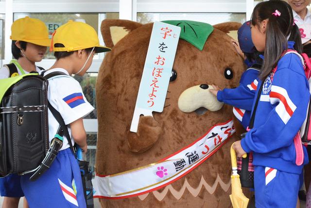 (写真)「字をおぼえよう」のプレートを手に持ち、登校した児童とあいさつするやはたいぬ