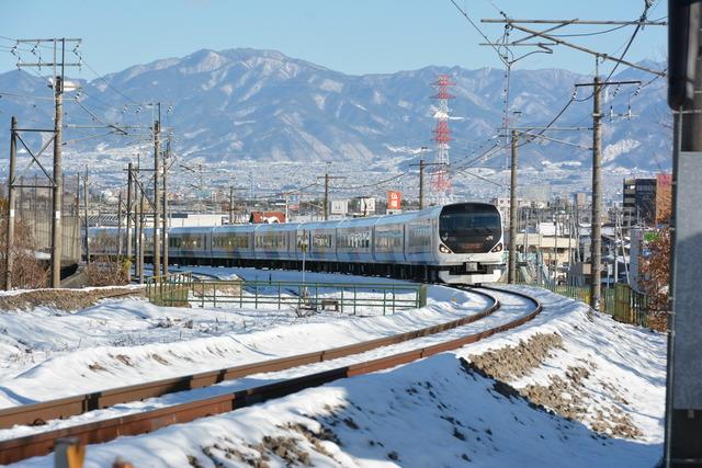 雪が積もった甲府盆地を背景に走行する白い車体の「E572系あずさ号」の写真