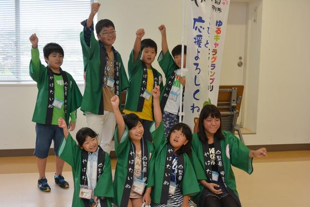 「やはたいぬ」と書かれた緑の法被を着た「やはたいぬ応援団」の子どもたち8人が片手を挙げている写真