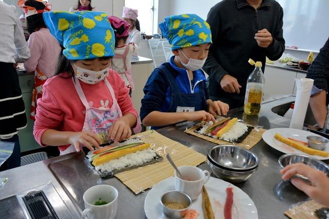 2人の子どもが酢飯の上に卵焼きやキュウリを乗せ恵方巻きを巻いている写真