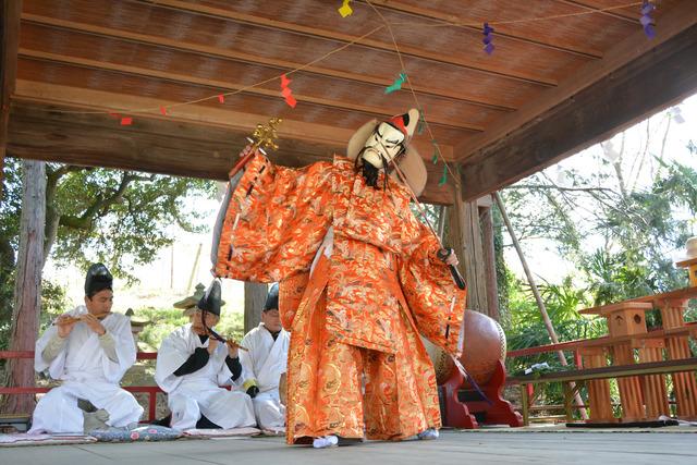 横笛の演奏に合わせてお面を付け橙の衣装を着た男性が神楽を舞っている写真