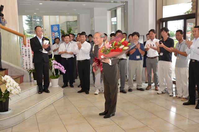 花束を手に持ち、多くの職員に拍手で迎えられる保坂市長の写真