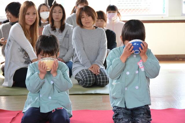 両手で茶碗を持ちお茶をいただいている園児とそれを見つめる保護者達の写真