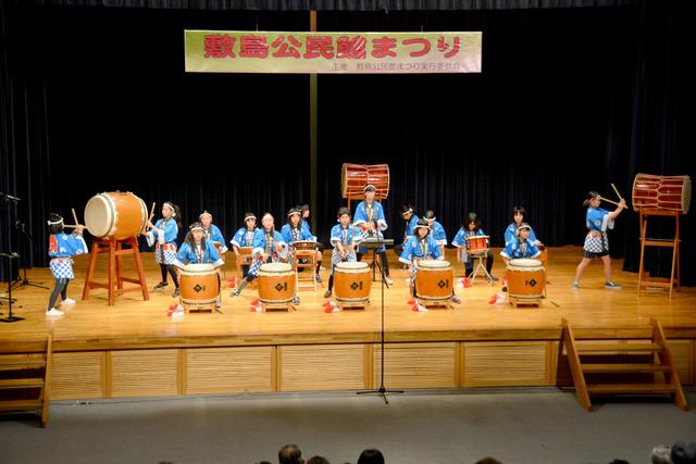 舞台の上で青い法被を着た小学生たちが和太鼓を叩いている写真