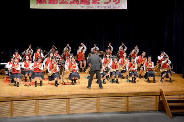 舞台の上で赤いベストを着た小学生たちが楽器の演奏をしている写真