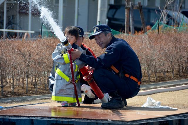 子ども用の消防服を着て、消防団の男性と共に放水を行う子どもの写真