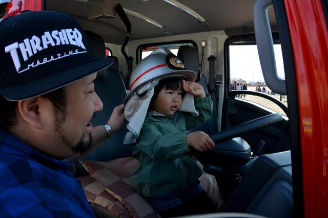 消防車両の運転席に座ってハンドルを握り、左手で敬礼をしている小さな子どもの写真