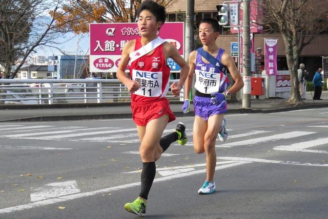 赤いユニフォームの甲斐市の選手と紫のユニフォームの甲府市の選手が僅差で走っている写真
