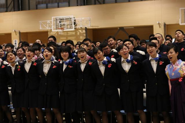 スーツを着た多くの卒業生と袴姿の先生が肩を組んで歌っている写真