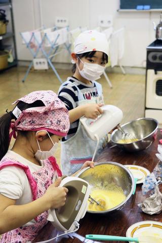 エプロン姿の小学生が泡だて器でケーキの生地を混ぜている様子の写真