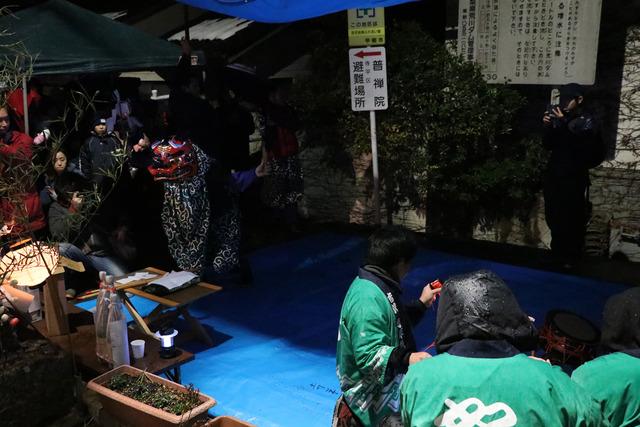 雨の中、緑の法被を着た男性が横笛や太鼓を演奏に合わせて獅子舞が披露されている写真