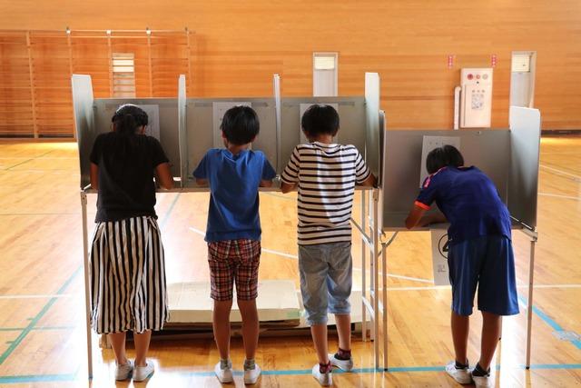 竜王西小学校の体育館にて6年生の児童四人が選挙の投票用紙に記入している写真