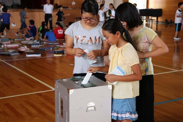 竜王西小学校の体育館にて6年生の女子児童三名が選挙の投票用紙を投票している写真