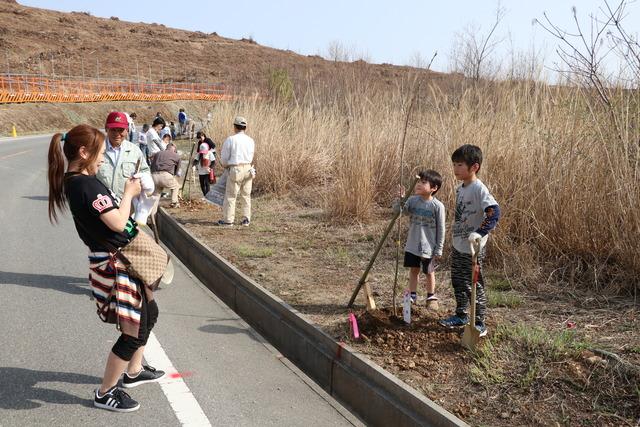 植樹した桜の前でポーズをとる二人の男の子とその姿を撮影する女性の写真